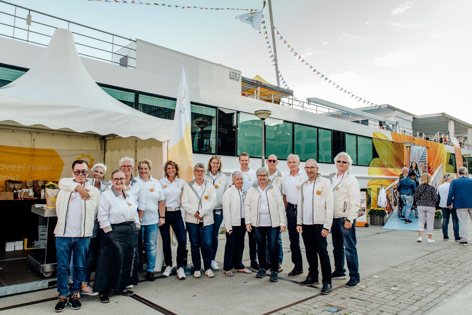 Een groep mensen in witte jassen poseert buiten voor een groot wit gebouw met gele accenten en een tent.