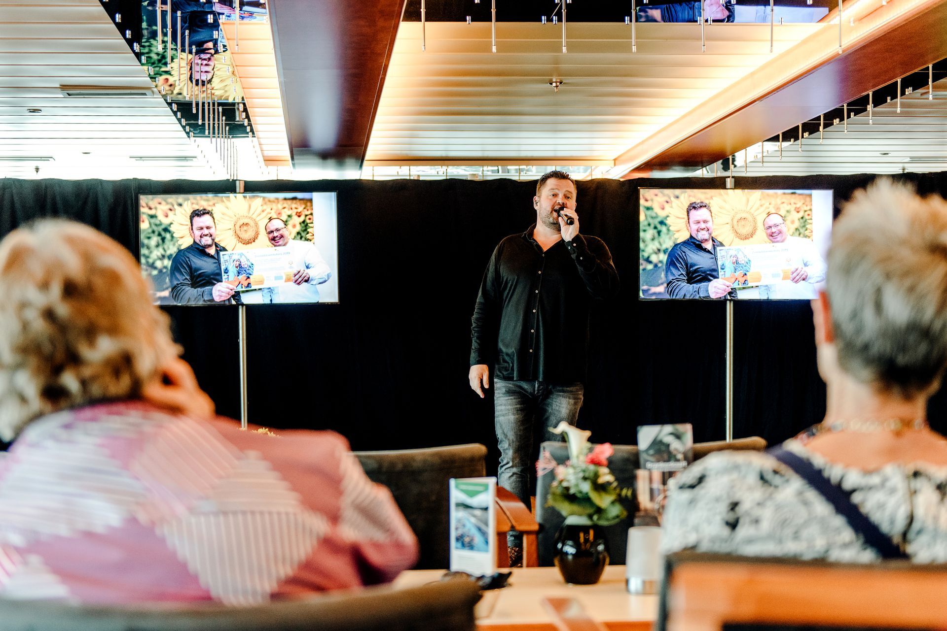 Man zingt op het podium, twee schermen tonen hetzelfde beeld. Twee toeschouwers zitten.