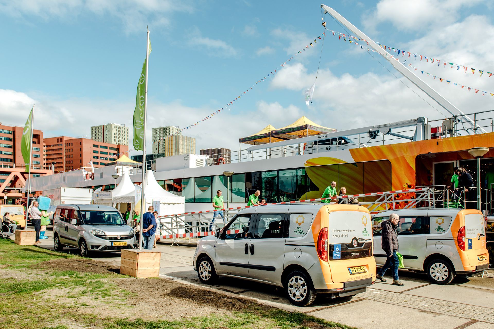 Drie busjes geparkeerd buiten een festivalterrein. Mensen in groene shirts, zonnige dag.