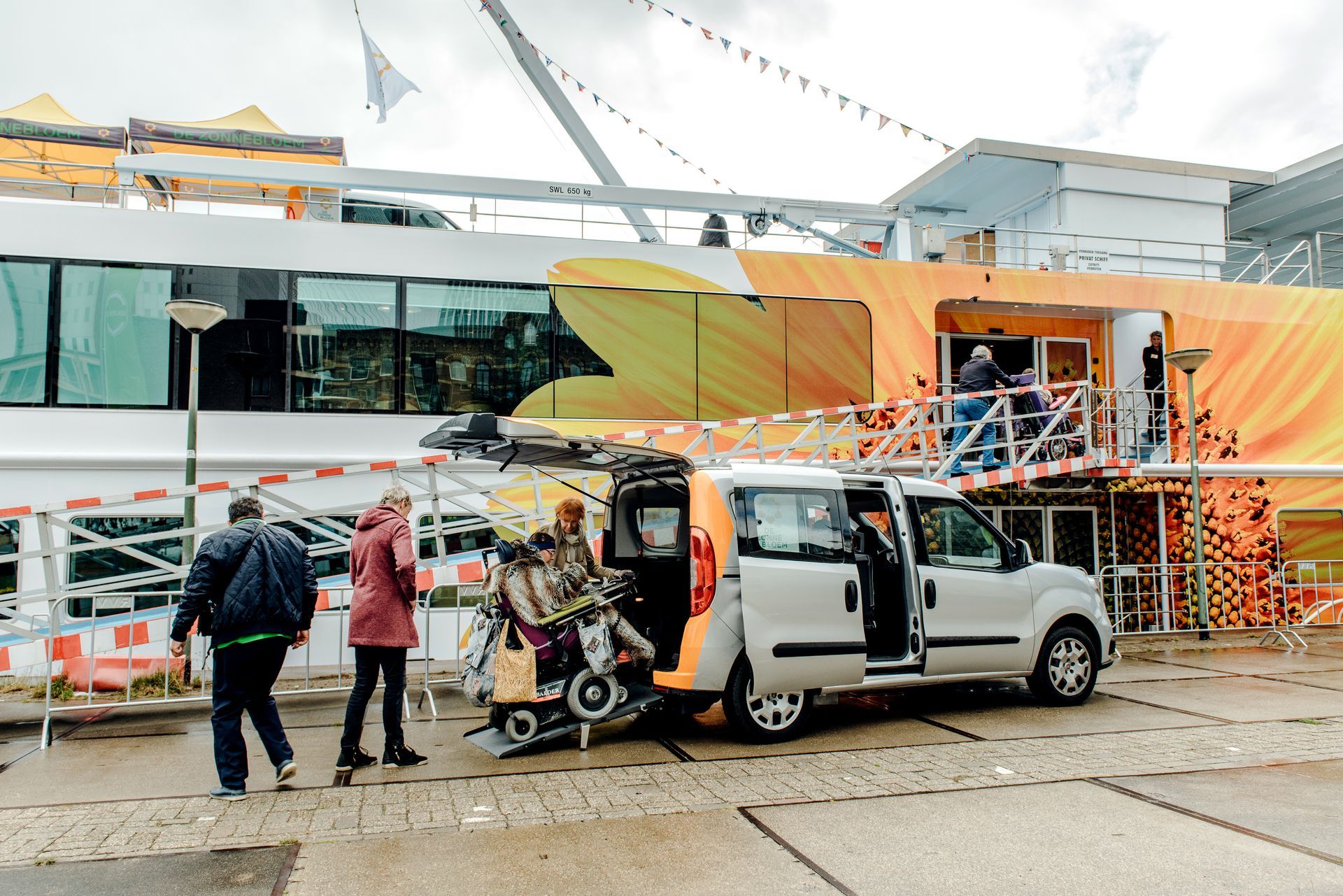 Een rolstoelbusje laadt een rolstoel uit bij een gebouw met een oranje en geel ontwerp. Mensen staan ernaast.