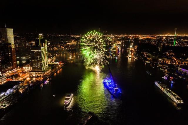 Vuurwerk boven een haven in een stad bij nacht, met verlichte gebouwen, boten en donker water.
