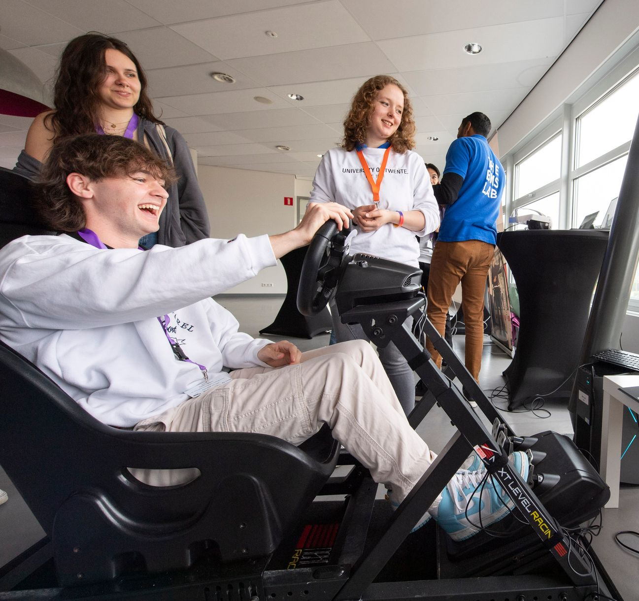 Persoon in een racesimulator, lachend, met twee mensen die toekijken; lichte binnenomgeving.