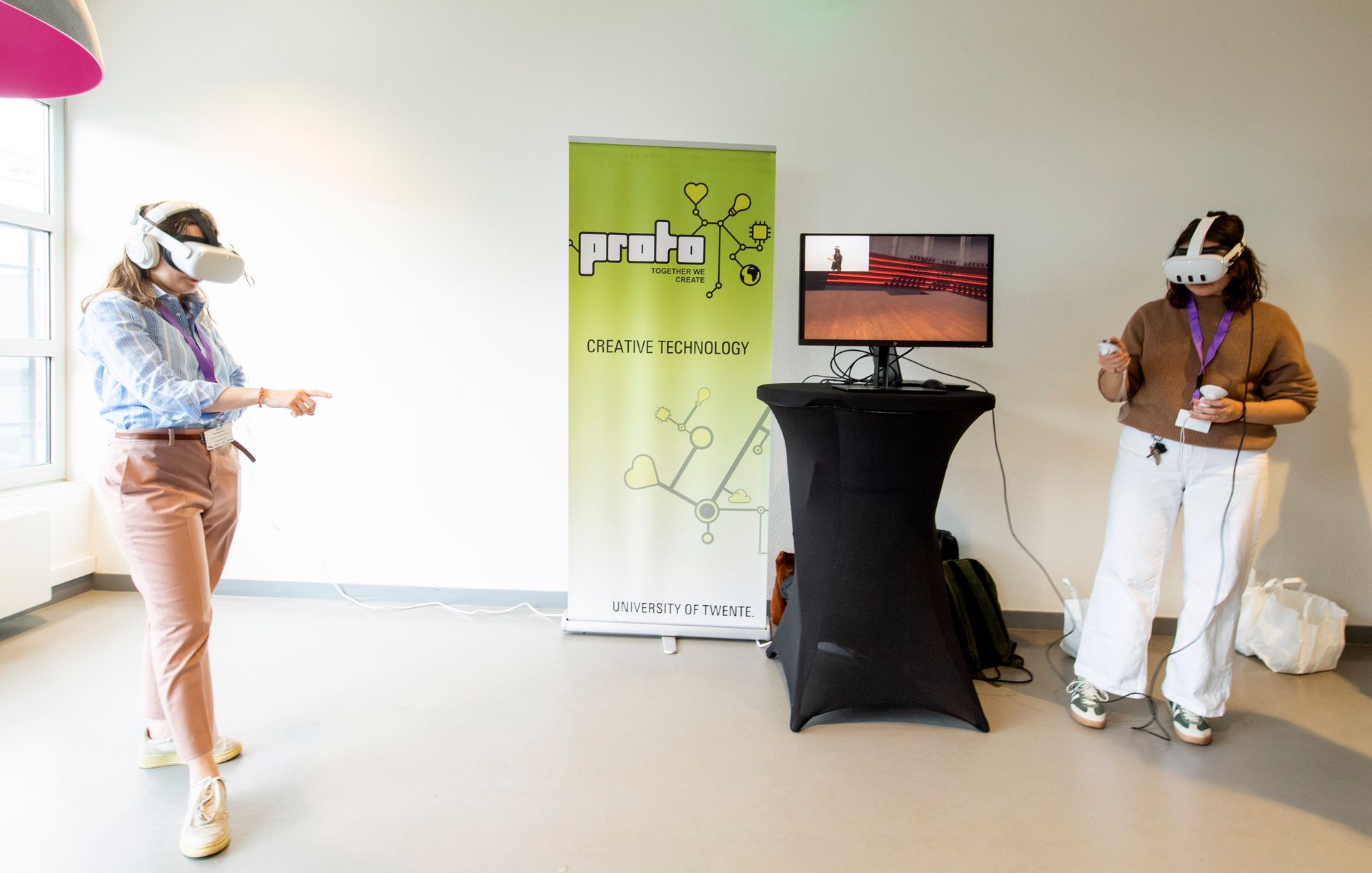 Twee mensen met een VR-bril interacteren met een virtuele omgeving; een scherm geeft hetzelfde beeld weer.