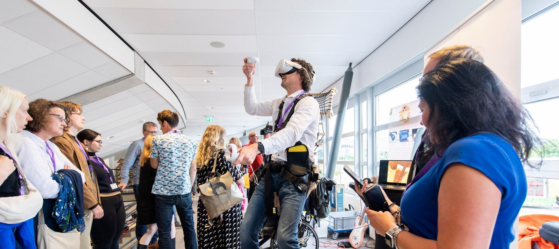 Persoon met VR-headset en controllers, omringd door toeschouwers. Vrouw met telefoon. In een goed verlicht gebouw.