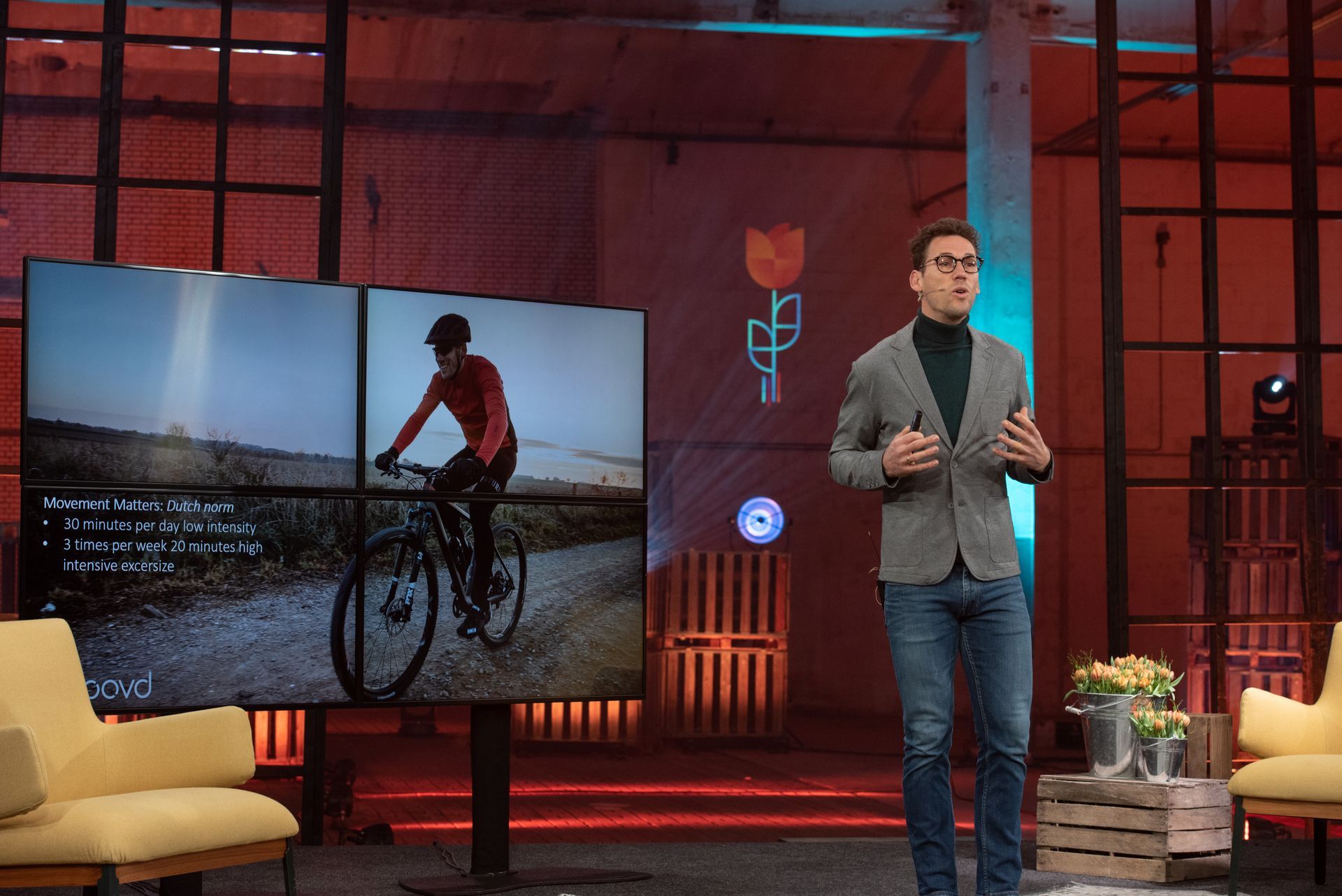 Man in blazer spreekt, gebarend naar scherm met fietser. Podium met schermen, logo en houten kratten.