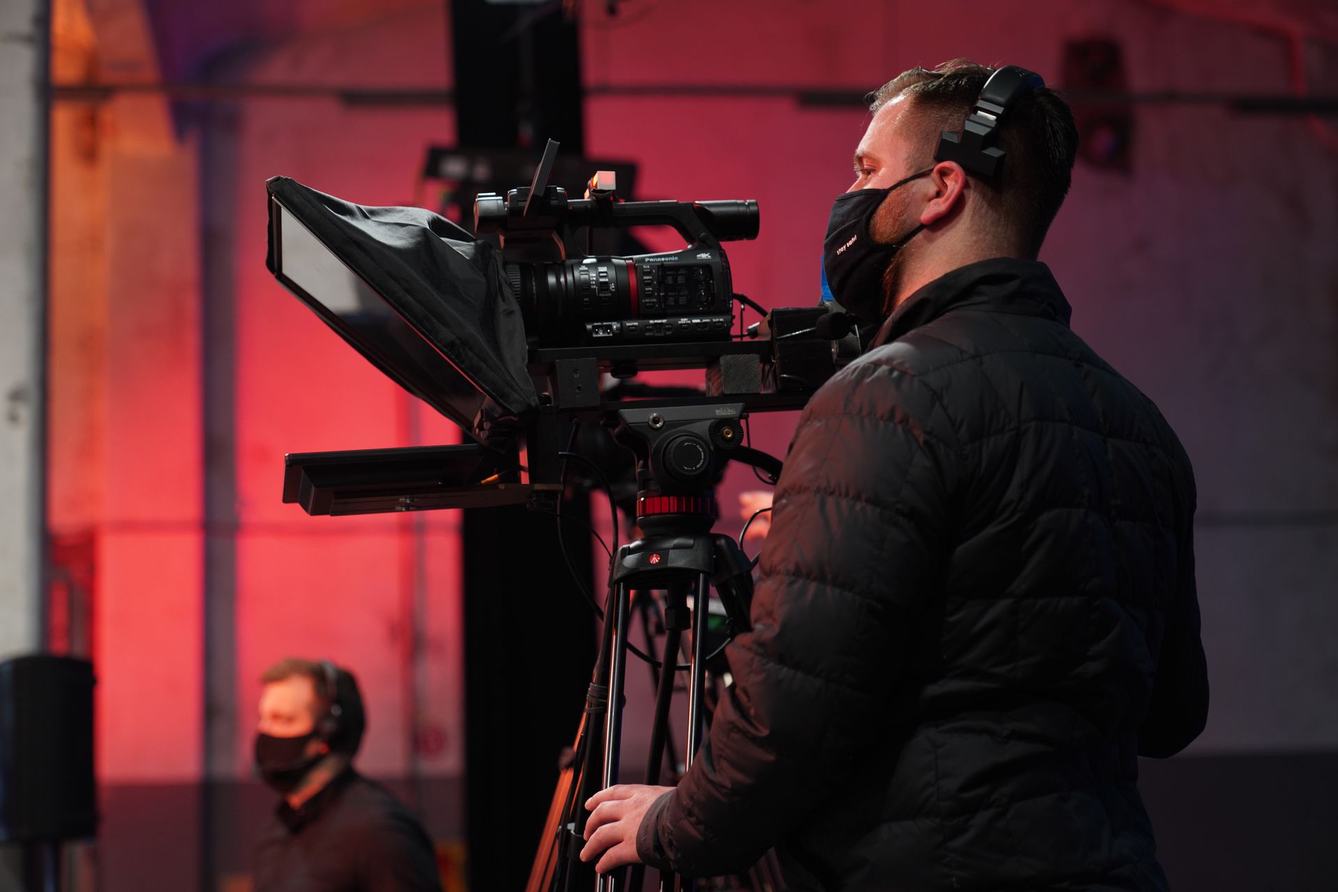 Een cameraman met een masker stelt de camera in op een statief in een studio met rode verlichting.