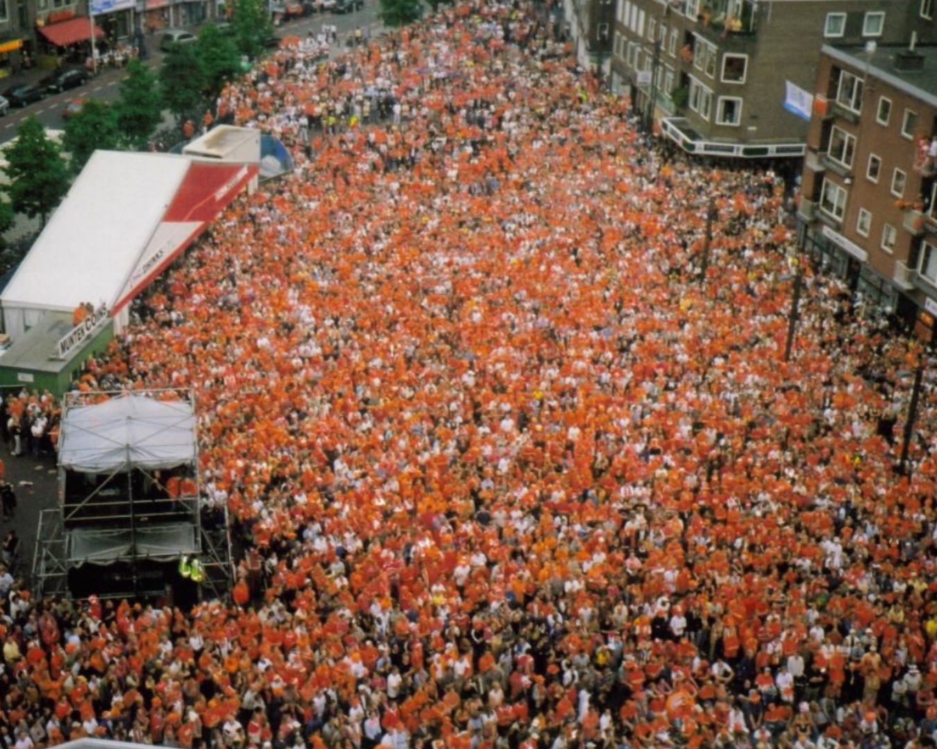 Een grote menigte gekleed in oranje kleding verzamelt zich op een stadsplein, waarschijnlijk om een ​​gebeurtenis te vieren.
