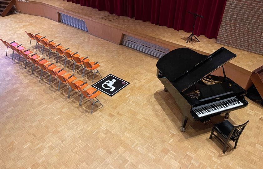 Hoogtebeeld van een theaterpodium met een vleugelpiano, zitplaatsen en een toegankelijkheidssymbool op de vloer.