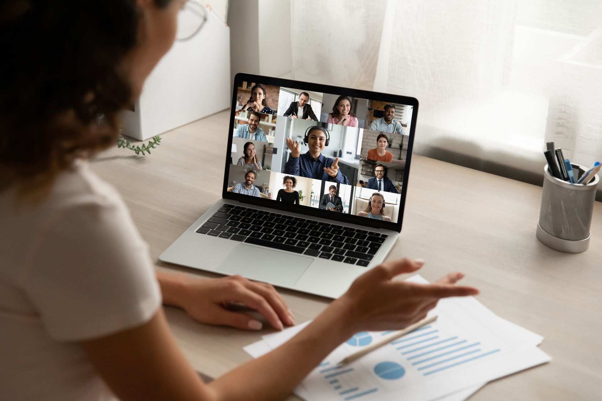 Woman in video call with colleagues, pointing to a document. Laptop screen shows multiple participants.