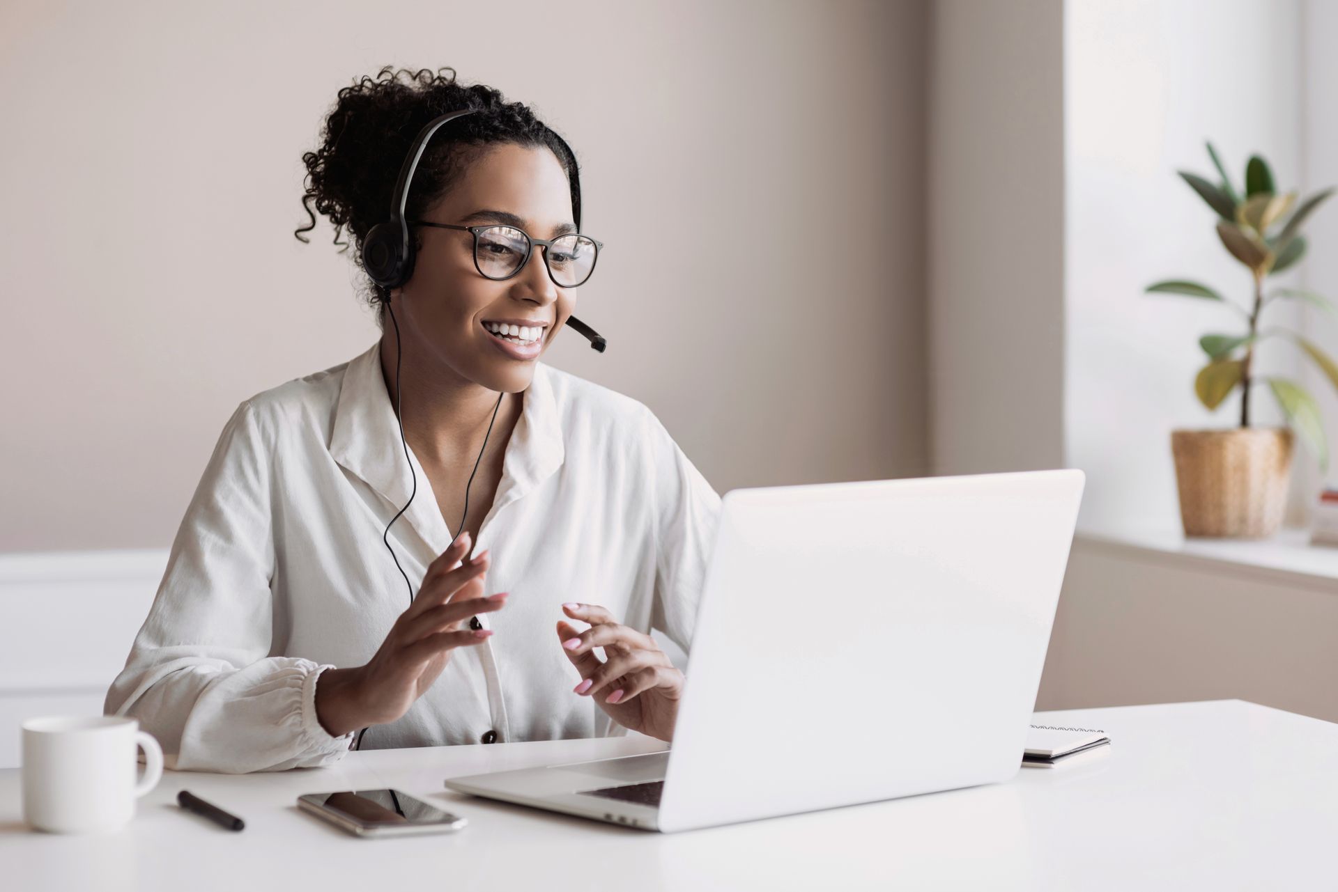 Woman wearing headset, smiles while gesturing at laptop screen, sitting at a desk indoors.