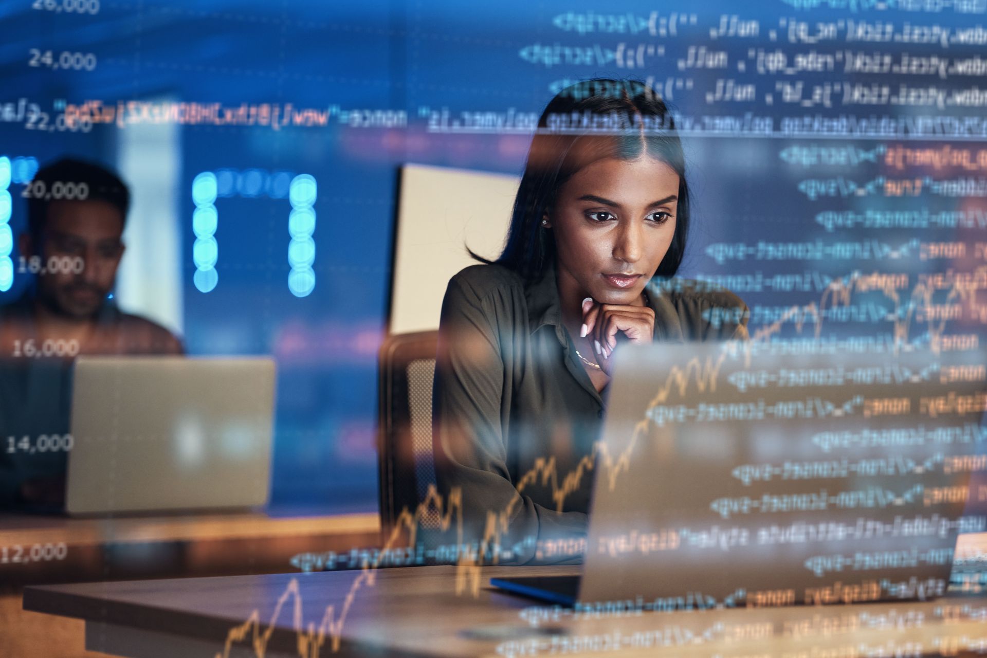 Woman focused on laptop, surrounded by data code in a dim office. Man working in the background.