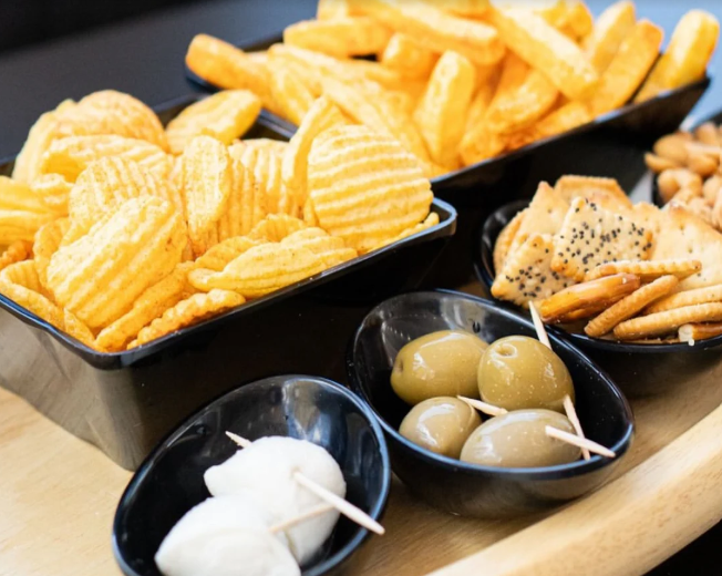 Assortimento di snack: patatine fritte, olive, cracker e mozzarella con stuzzicadenti, serviti in ciotole nere su un vassoio.