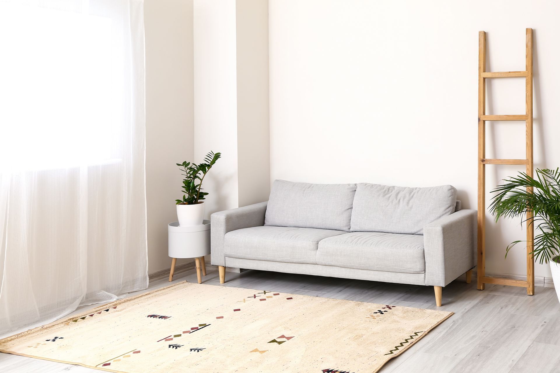 Neutral-toned living room with a gray sofa, rug, ladder, and plants near a window with sheer curtains.
