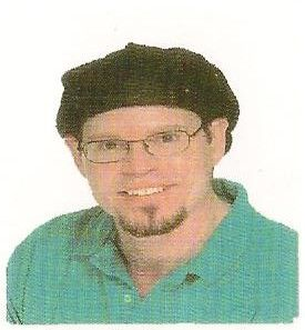 Man with glasses, goatee, and black cap smiles, wearing a green collared shirt.