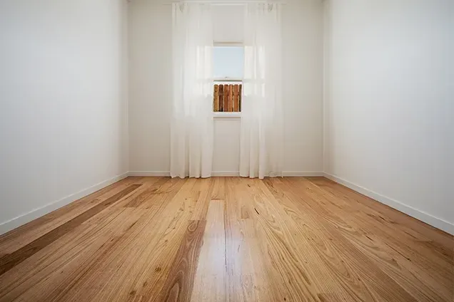 Empty room with wood floors, white walls, and sheer curtains over a window.