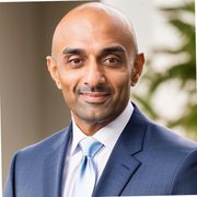 Headshot of a smiling person wearing a blue suit, white collared shirt, and light blue tie against a soft-focus background.