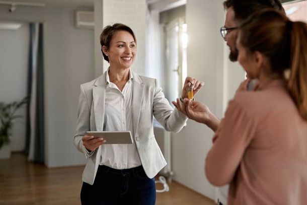 A smiling real estate agent hands a set of keys to a client inside a bright, unfurnished room.