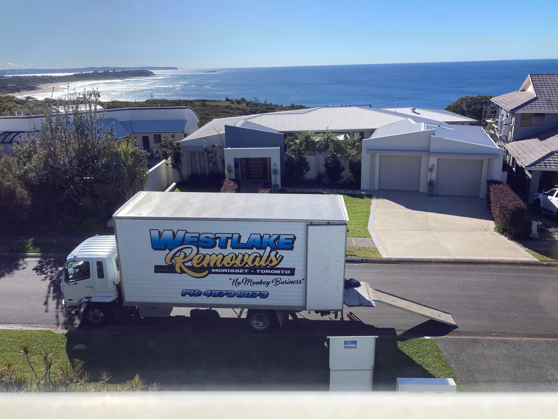 Moving truck parked in front of a house overlooking a beach; blue ocean and sky in background.