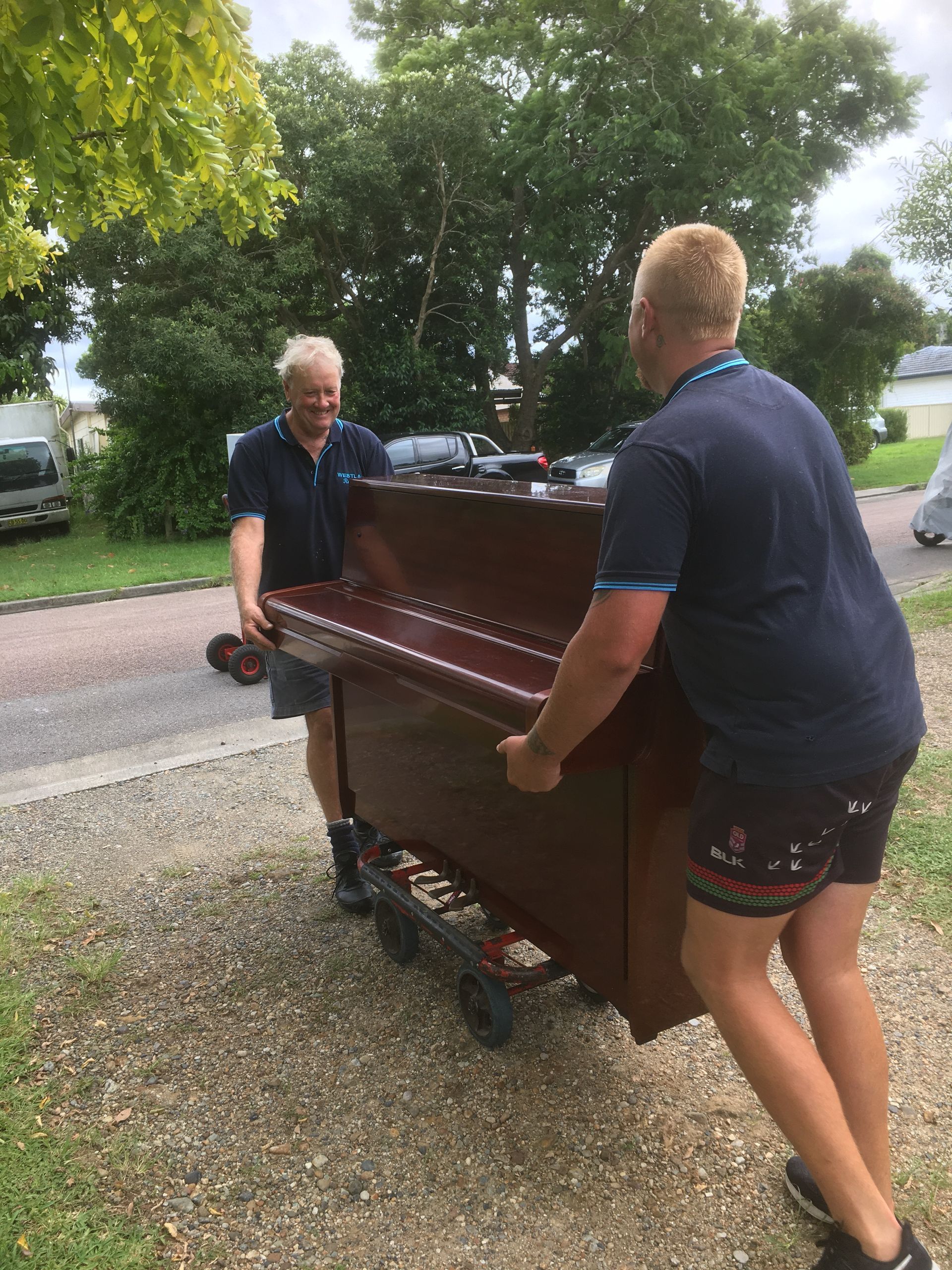 Two men moving a brown piano on a wheeled dolly along a gravel driveway outdoors.
