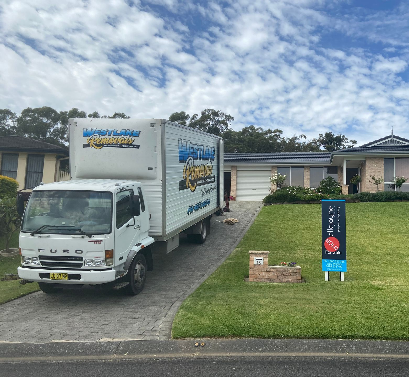 White moving truck driving on a paved road through a rural area under a blue sky.