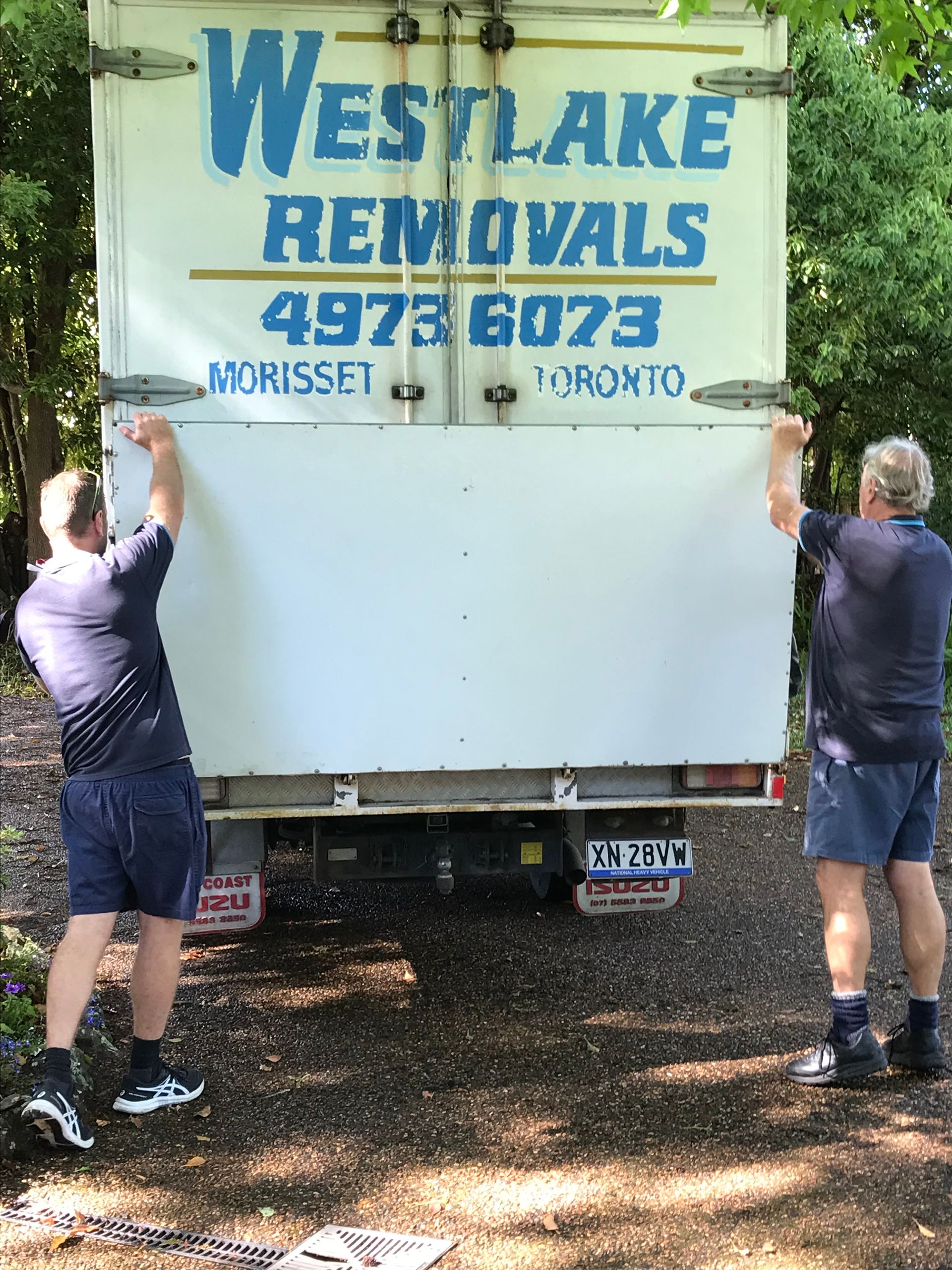 Two men lifting the rear door of a Westlake Removals truck in a wooded area.