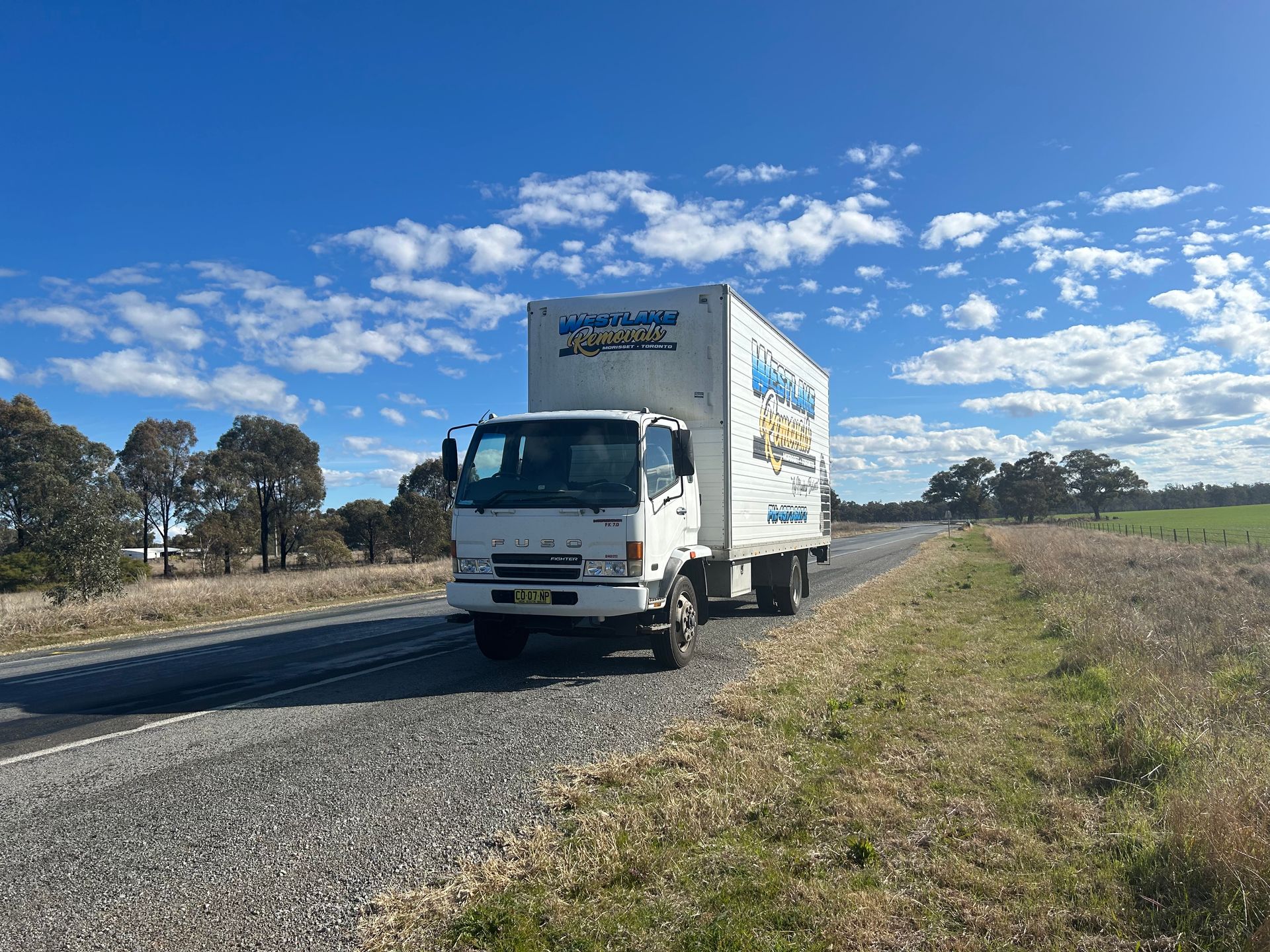 White truck driving on a rural road under a blue sky with clouds.