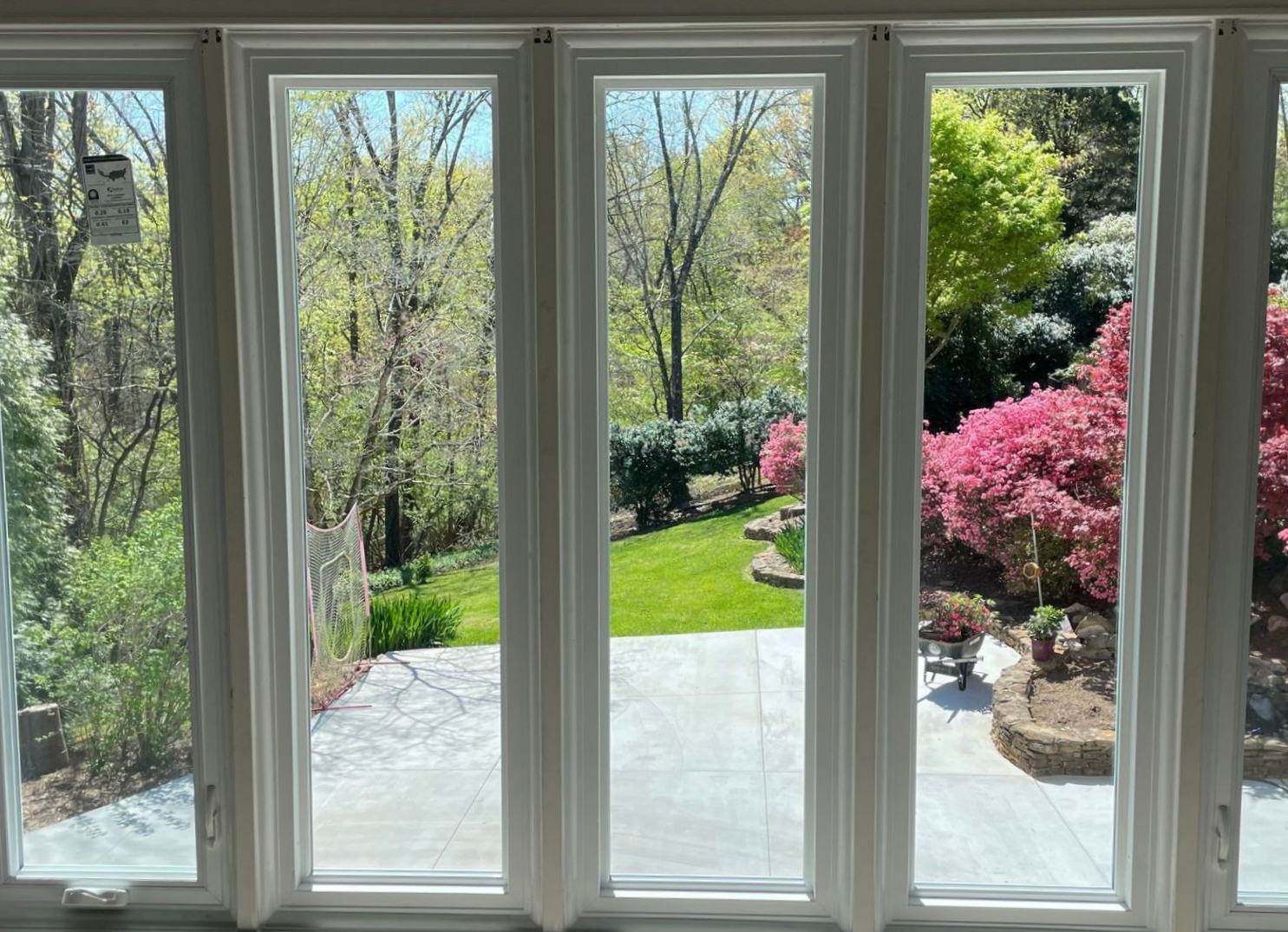 A multi-pane bay window looks out onto a sunny backyard with a concrete patio, green lawn, and flowering pink bushes.
