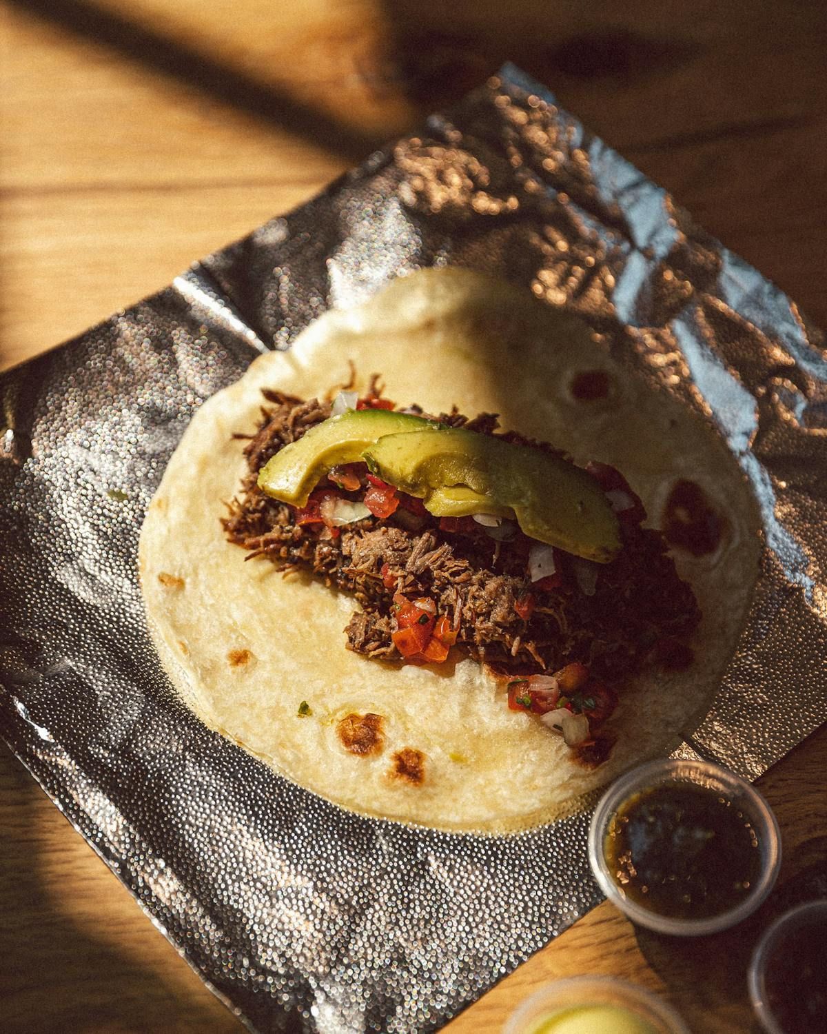 A flour tortilla taco topped with shredded meat, diced tomatoes, onion, and sliced avocado, sitting on crinkled foil.