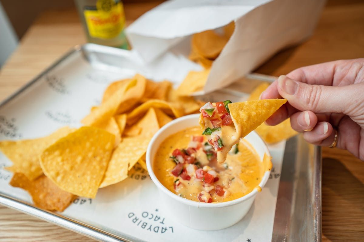 A hand holds a tortilla chip dipped into a small cup of queso with pico de gallo, beside a tray of more chips.