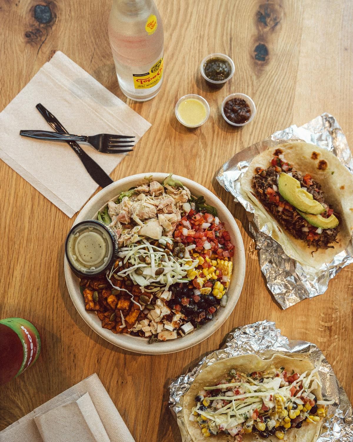 Three breakfast tacos on a metal tray with a cup of coffee, a latte, and three small containers of dipping sauces.