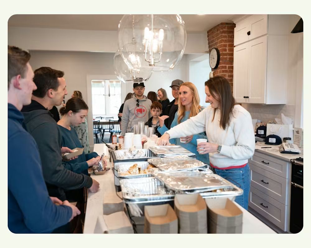 A line of people stands at a kitchen counter being served food by two individuals in a bright, modern, home-like setting.