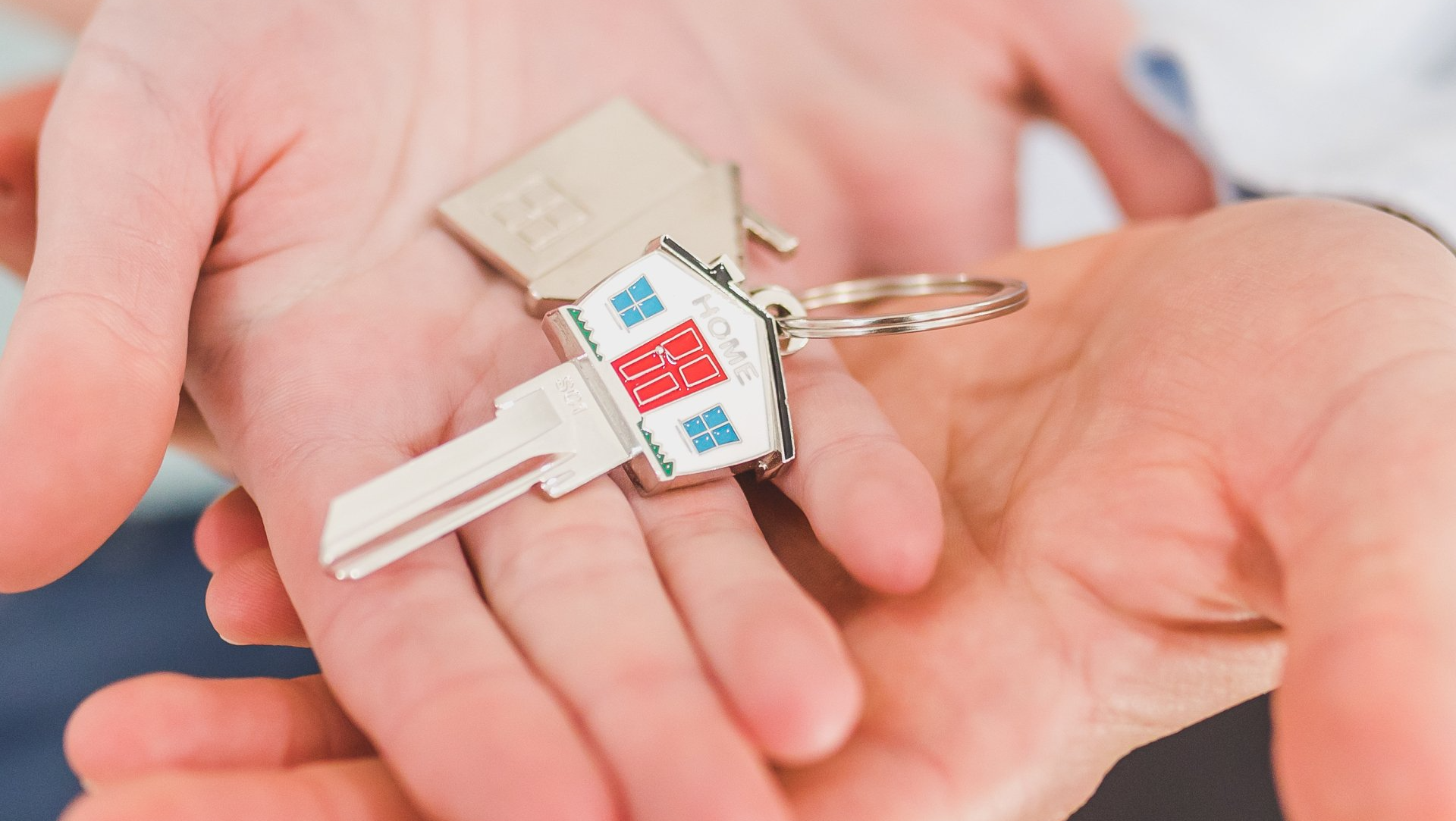 Two hands holding house keys on a keychain, one key shaped as a house with a red roof.