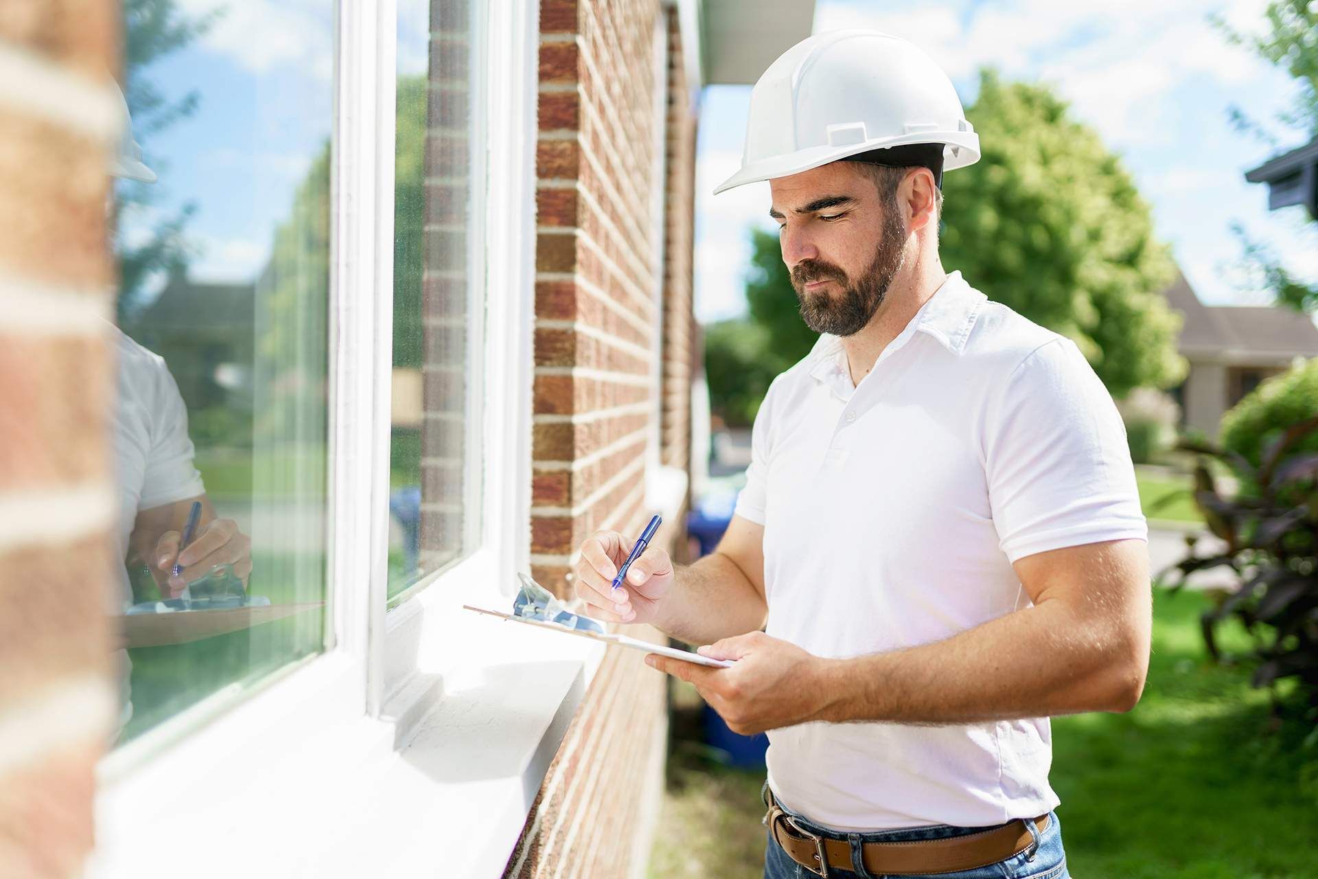 a man wearing a hard hat is looking at a window while holding a clipboard .