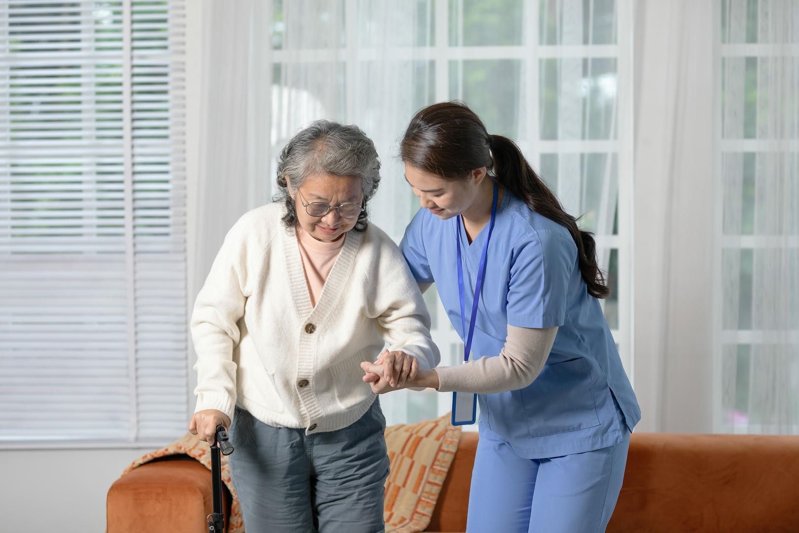 A nurse is helping an elderly woman walk in a living room.