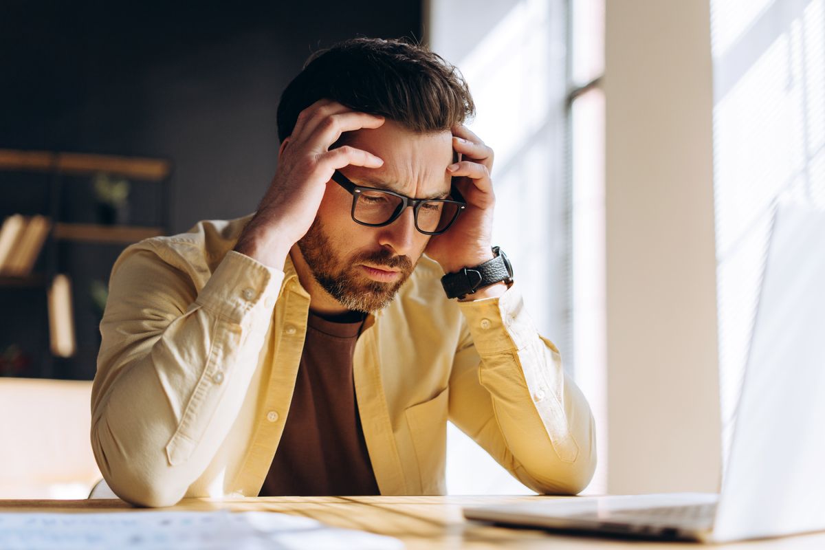 Man with glasses holding head, appears stressed while sitting at a desk with a laptop.