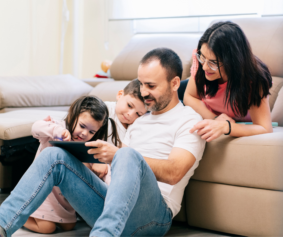 Family of four looking at a tablet together on the floor in front of a couch; smiles.