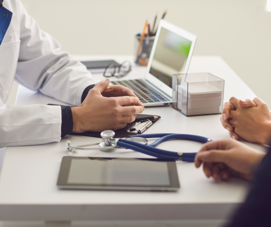 A doctor and patient sitting across from each other at a desk with a laptop, tablet, and stethoscope.