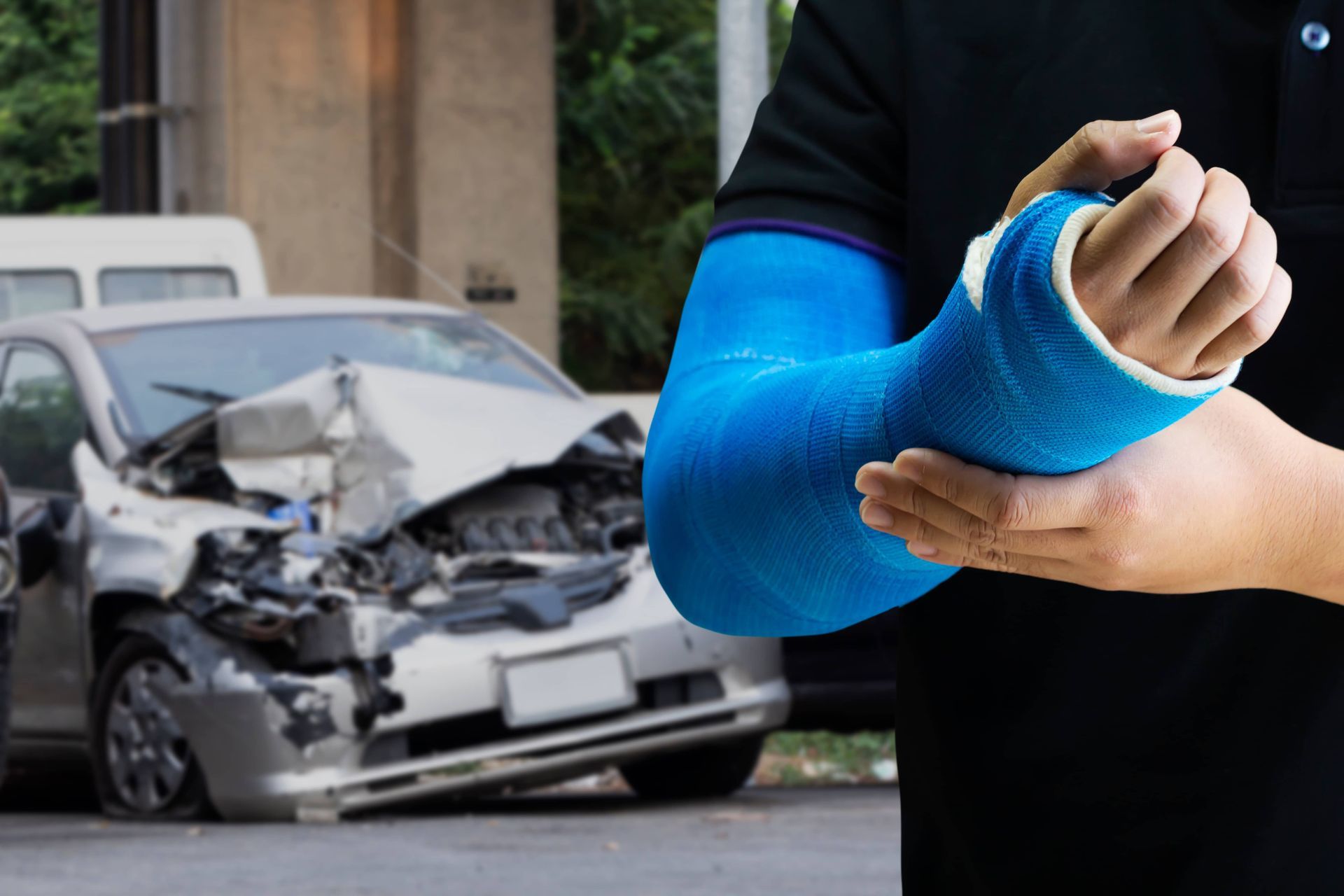 Person with blue cast on arm in foreground, with a badly damaged car after a crash in the background.