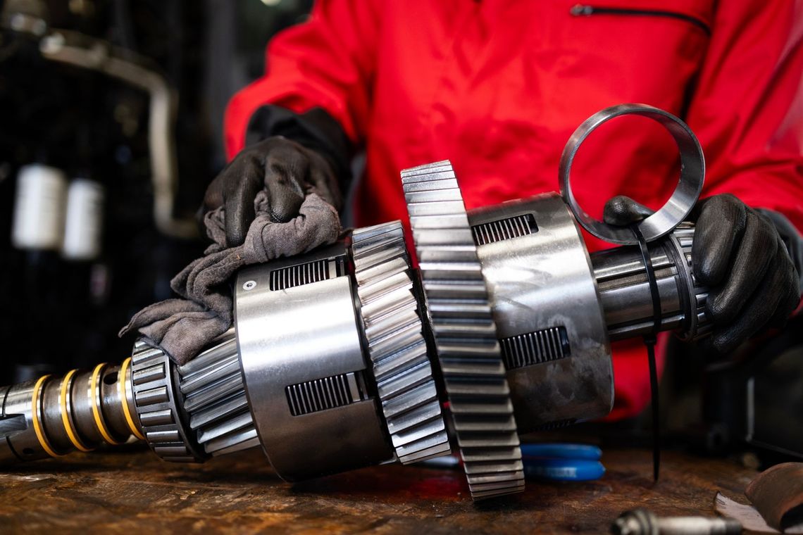 Hands in gloves inspecting a metal gear assembly on a workbench, with a red jacket in the background