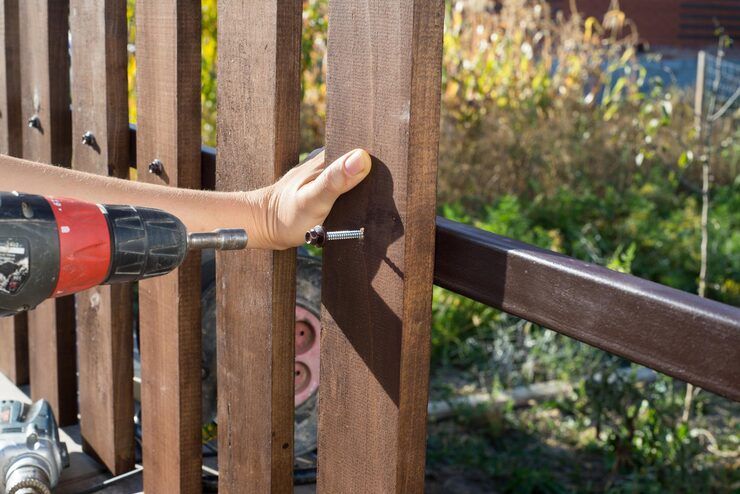 A person using a drill to attach a wooden post to a fence on a sunny day.