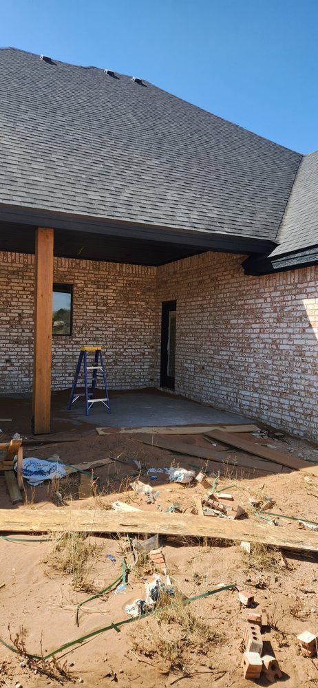 Brick home under construction, with a shaded patio, on a sunny day. Dirt ground and roofing visible.