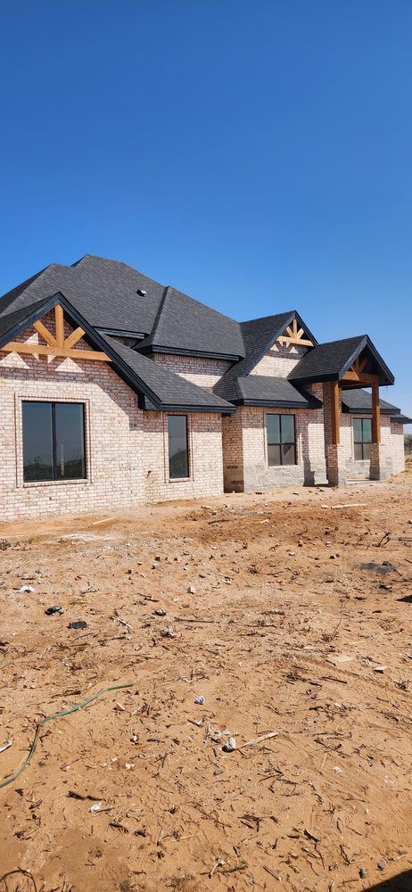 A newly constructed beige brick house with black roof under a clear blue sky. The yard is dirt.