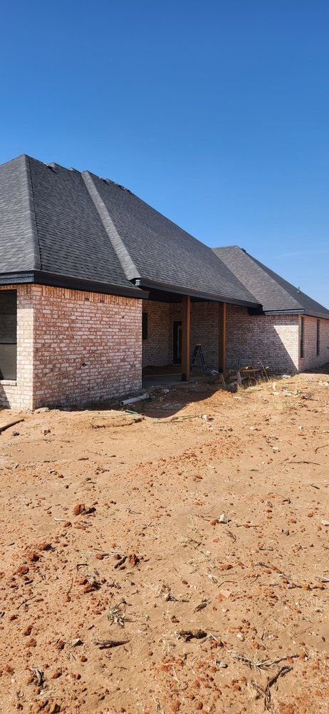Brick house under construction with black roof and exposed dirt. Bright blue sky overhead.