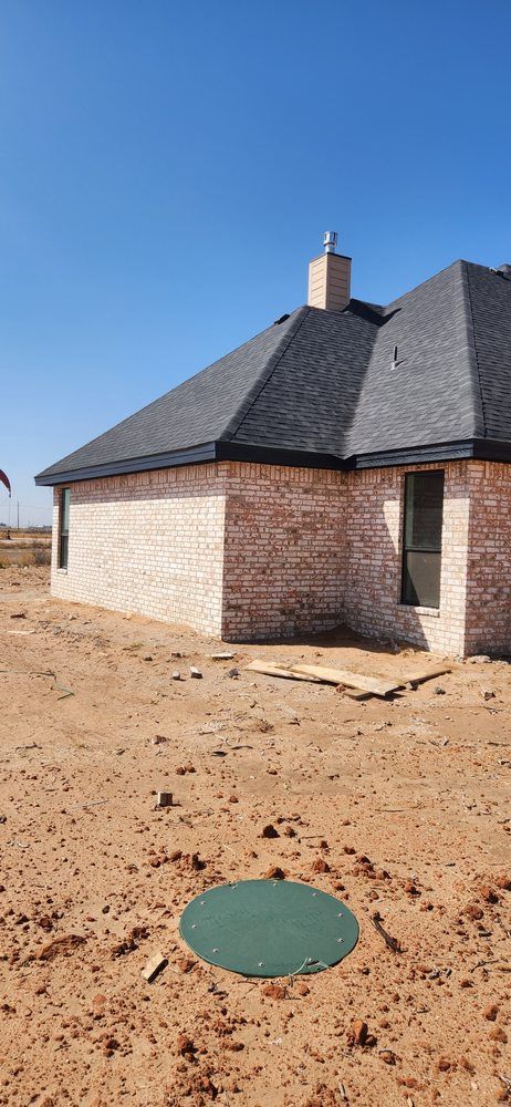 Brick house under construction on a dirt lot. Dark shingled roof and a green lid. Bright blue sky.