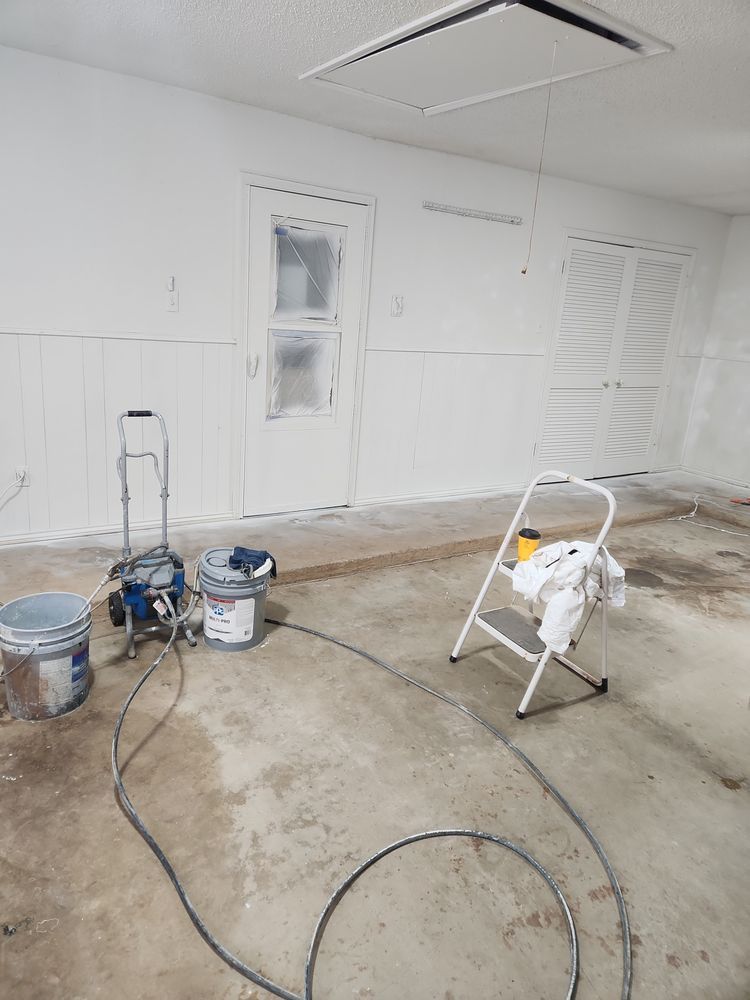 White-painted room interior in progress, paint sprayer, buckets, ladder, and taped-up doors.