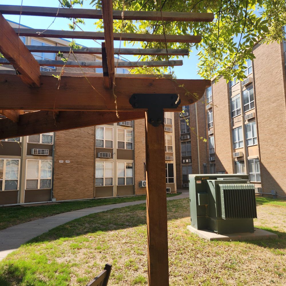 Wooden pergola in a grassy courtyard, with apartment buildings in the background and an electrical box.