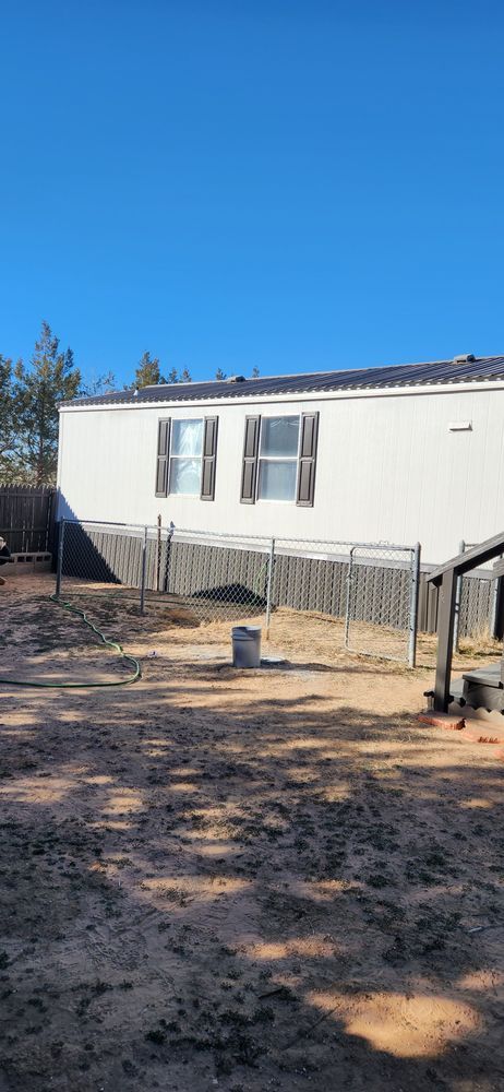 White house with blue sky. Backyard, fence, dirt ground, two windows with shutters.