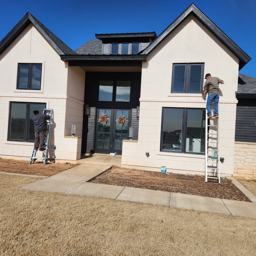 Two men cleaning windows on a two-story beige house with black trim under a clear blue sky.