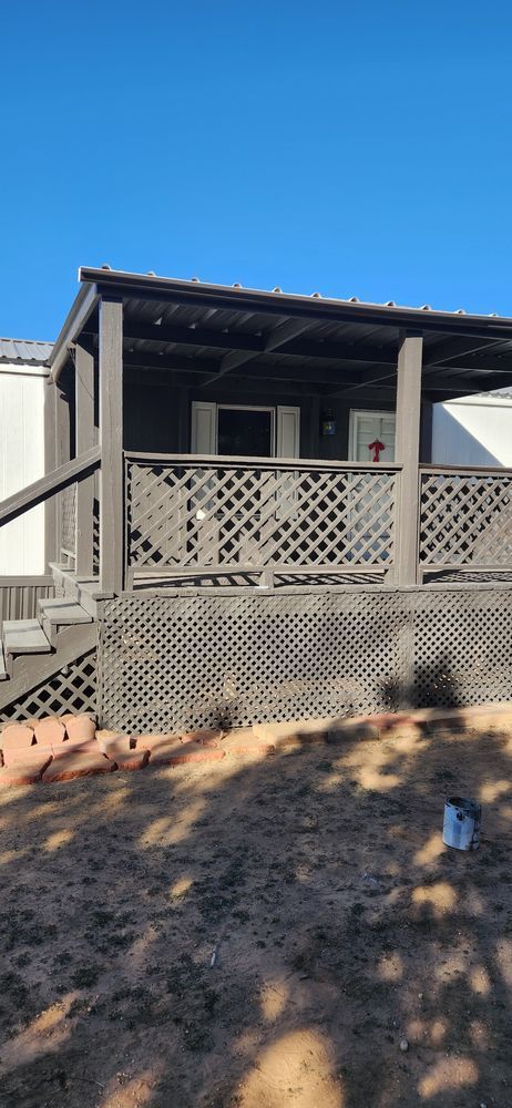 A brown house with lattice detailing and covered porch sits on a dirt lot. Blue sky overhead.