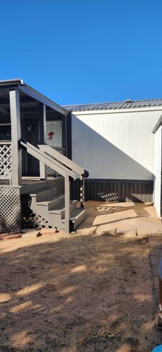 A mobile home with gray stairs and a porch. The yard is dirt, and the sky is blue.